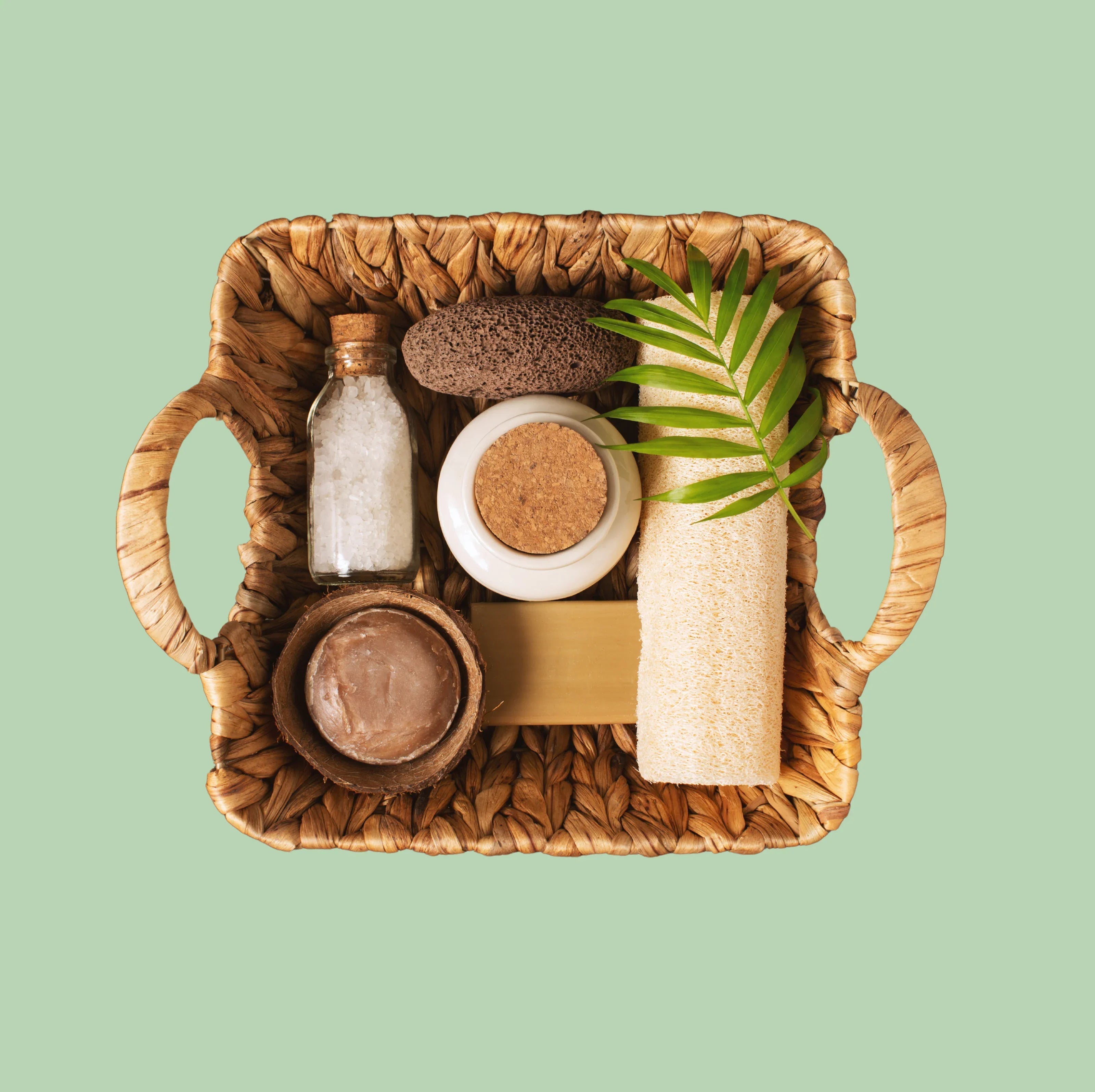 Wicker basket with natural wellness products, spa salt, soap, loofah, and leaf on green background